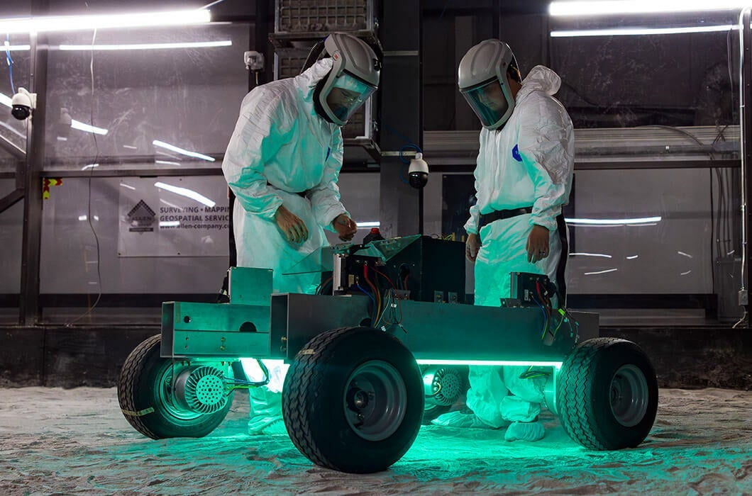 Two researchers wearing protective gear at UCF’s Exolith Lab examine a rover in the lab’s regolith bin — the world’s largest simulated lunar surface.