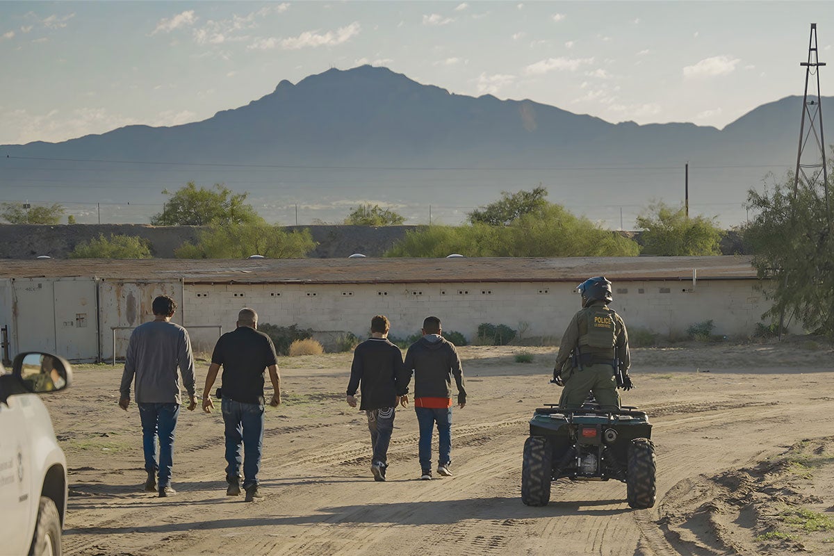 Four people walking as a border patrol agent rides an ATV beside them