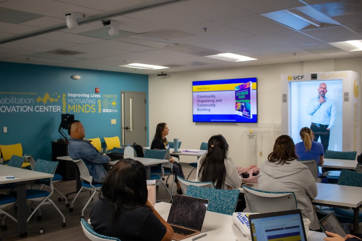 Students working at desks while listening to a hologram of a person speaking.