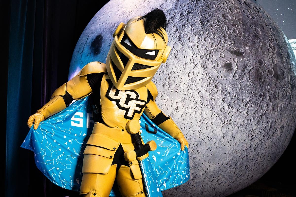 Knightro poses in front of a giant moon backdrop while wearing a blue, constellation-themed SpaceU cape