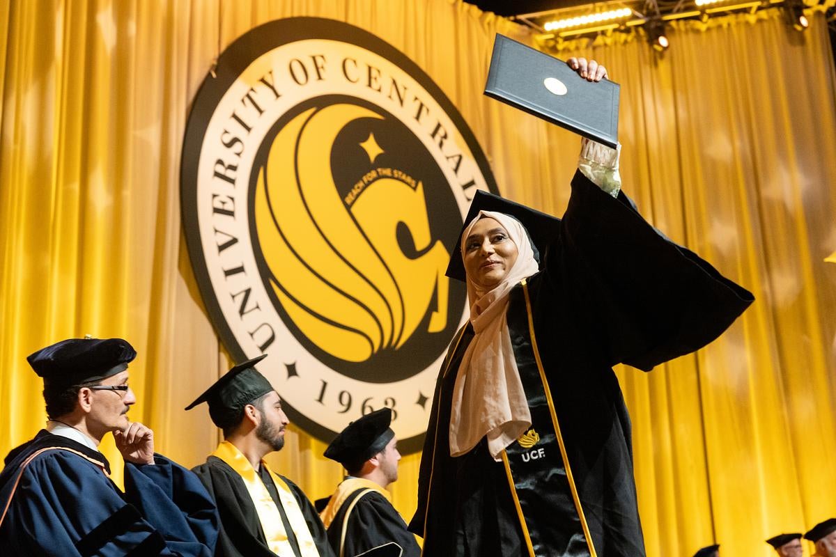 A graduate holds up their diploma while walking across the stage during UCF's Spring 2024 commencement ceremony at Addition Financial Arena