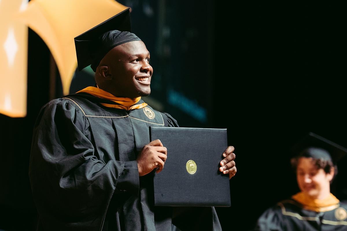 A graduate walks across the stage holding his diploma during UCF's Summer 2024 commencement ceremony at Addition Financial Arena