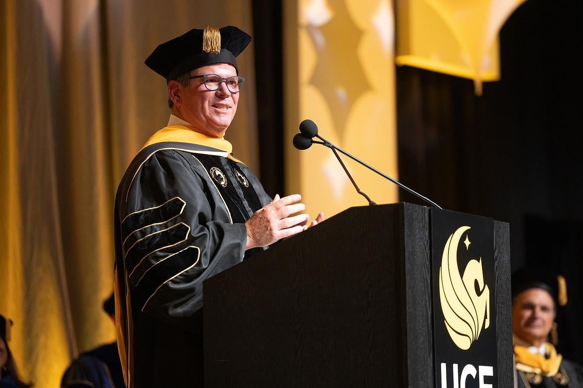 Ken robinson wearing a cap and gown and speaking at a commencement podium