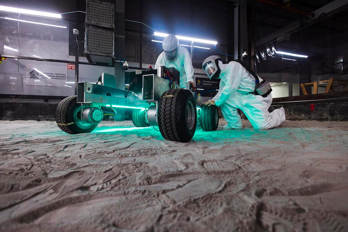 Two people wearing Hazmat suits operate a rover in the regolith bin at UCF's Exolith Lab