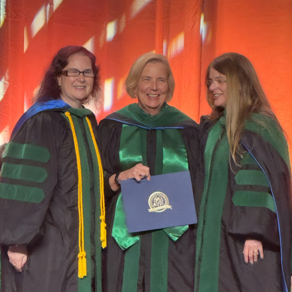 three people wearing commencement regalia and standing on stage