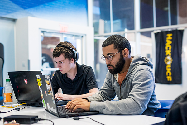 two students sitting next to each other. Each using their laptop.